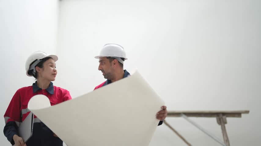 Two construction workers hold large blueprint while discussing building layout and reviewing project details together inside unfinished construction site.