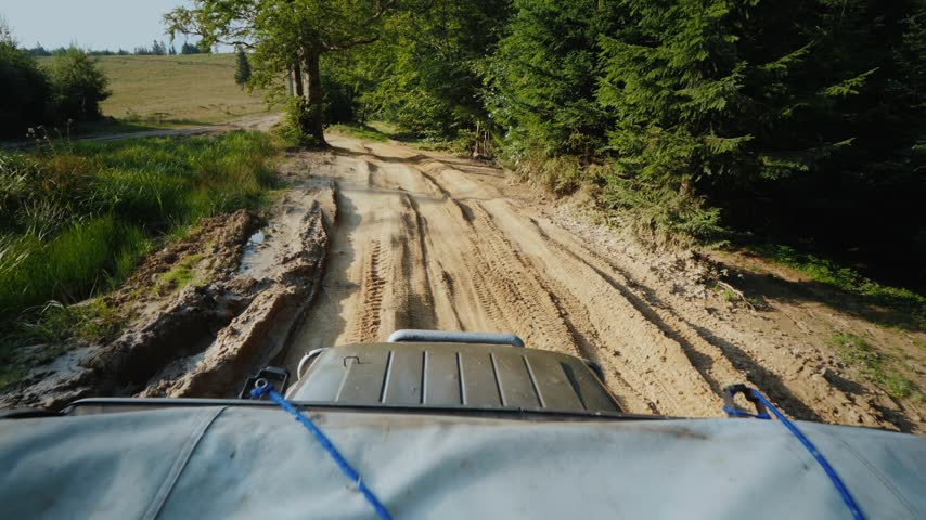 POV footage of 4x4 car driving over rugged dirt path in woods. Offroad adventure vehicle moves through muddy rutted road with lush green trees and fields on sunny summer day.