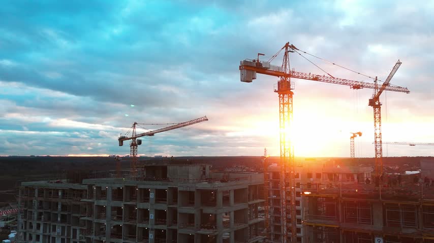 Sunset paints sky above busy construction site. Multiple cranes loom over unfinished buildings. Warm light highlights steel structures against clouds