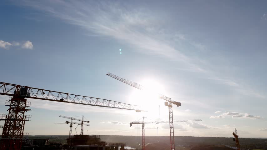 Construction cranes stand tall against a bright sky. Sunlight glints off metal structures as shadows stretch. Silhouettes of machinery hint at progress and industry