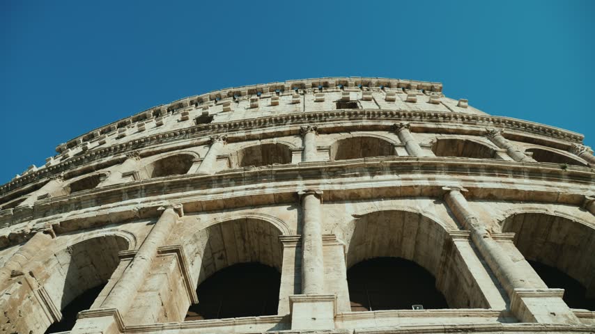 Low angle view captures iconic ancient Colosseum exterior in Rome Italy against clear blue sky. Cinematic travel clip features architectural details of famous historical landmark site.