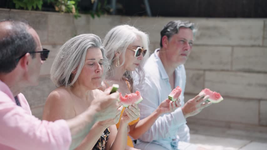 Four mature friends sitting together at the poolside on a sunny day. Joyful seniors laughing, talking, and enjoying fresh watermelon slices