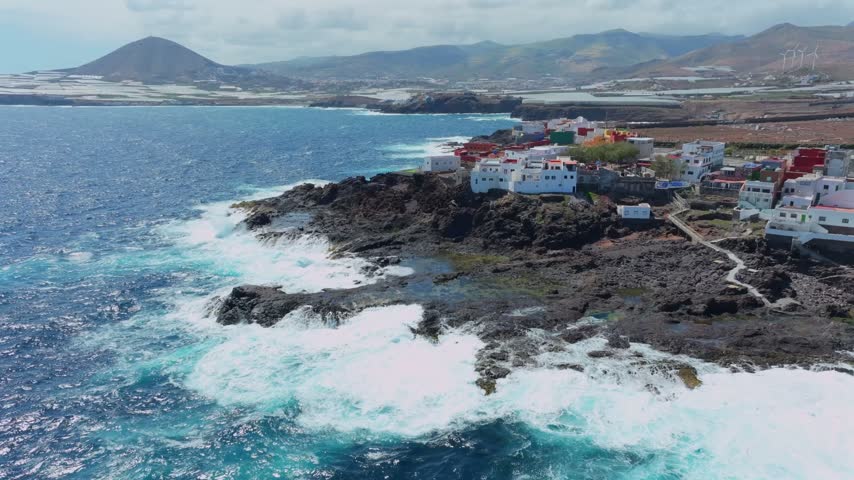 Powerful ocean waves crashing against the rugged volcanic coastline of Punta de Galdar. Small coastal village with white houses on Gran Canaria