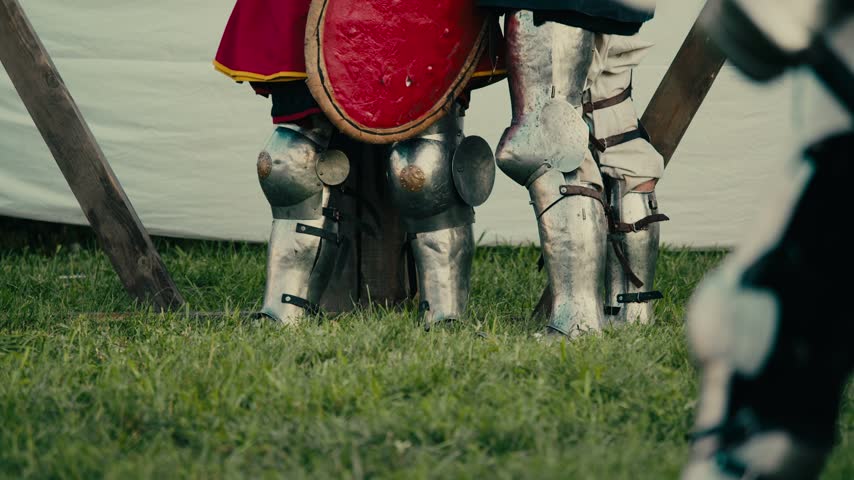 Close up highlights steel leg armor on standing knights. Detailed metal greaves protect warriors waiting for brutal historical combat tournament on grassy field