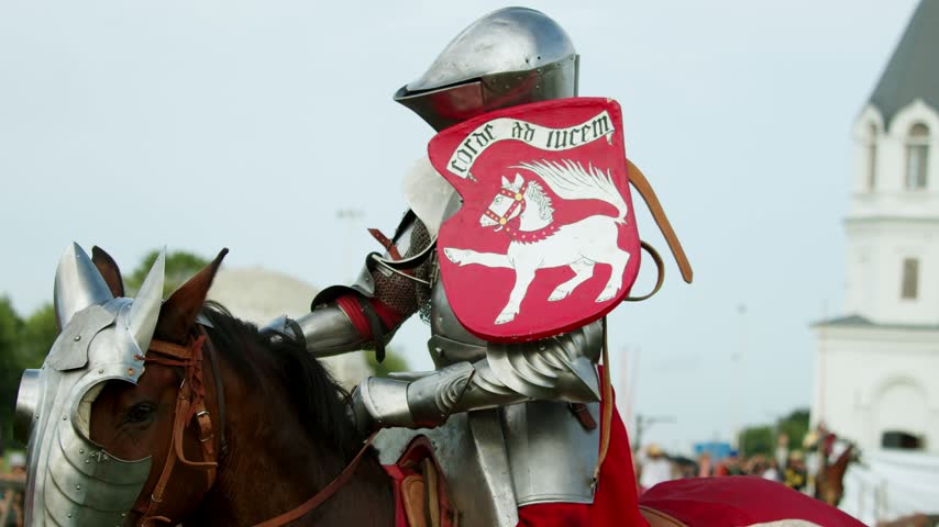 Helper hands striped wooden lance to mounted knight in heavy armor. Fully geared medieval rider sits on armored horse preparing for authentic jousting tournament in front of crowd