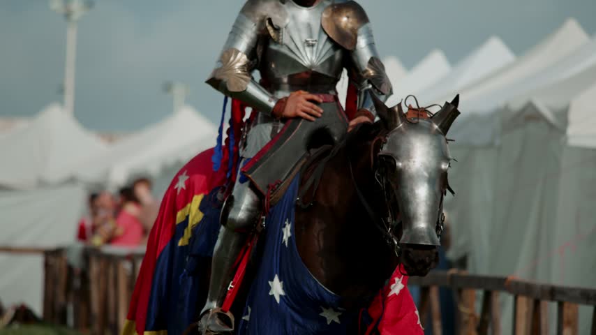 Armored knight rides decorated warhorse past white festival tents. Metal chamfron protects animal head while heavy cavalry soldier moves toward medieval jousting tournament arena