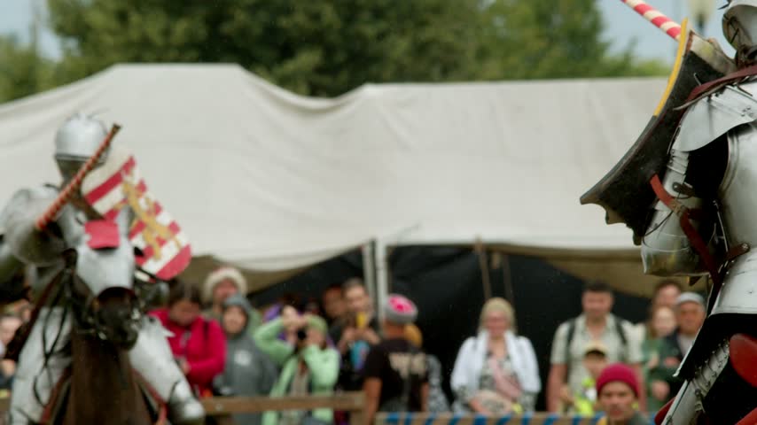 Long wooden lance misses rival shield during rainy jousting clash. Blurred crowd watches medieval knights fighting on horseback near white festival tents in bad weather