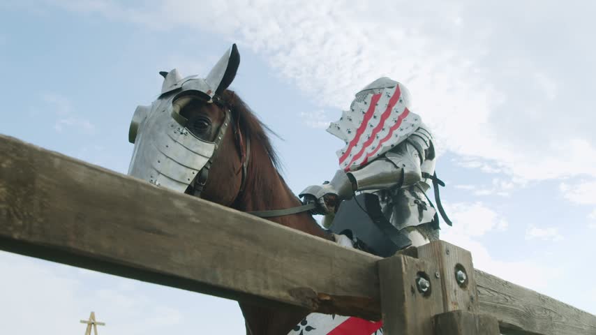 Armored knight on horseback rides slowly along wooden fence. Low angle shot captures shining metal gear and decorated shield against cloudy blue sky background during medieval historical festival