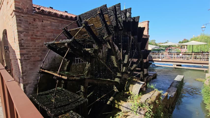 Historic Moss-Covered Waterwheel At Old Brick Mill Along A Sunny Canal With Rustic Charm