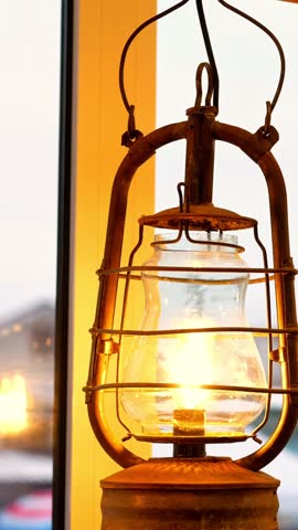 Old rusty kerosene lantern burning bright near window glass frame surface view. Retro metal oil lamp emitting warm golden fire light illumination inside rustic country house room habitat.