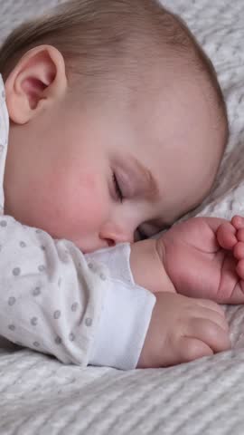 Peaceful little newborn child sleeping soundly on white textured blanket surface. Sweet tiny toddler wearing polka dot pajamas resting in bed during healthy daytime nap at cozy home nursery.