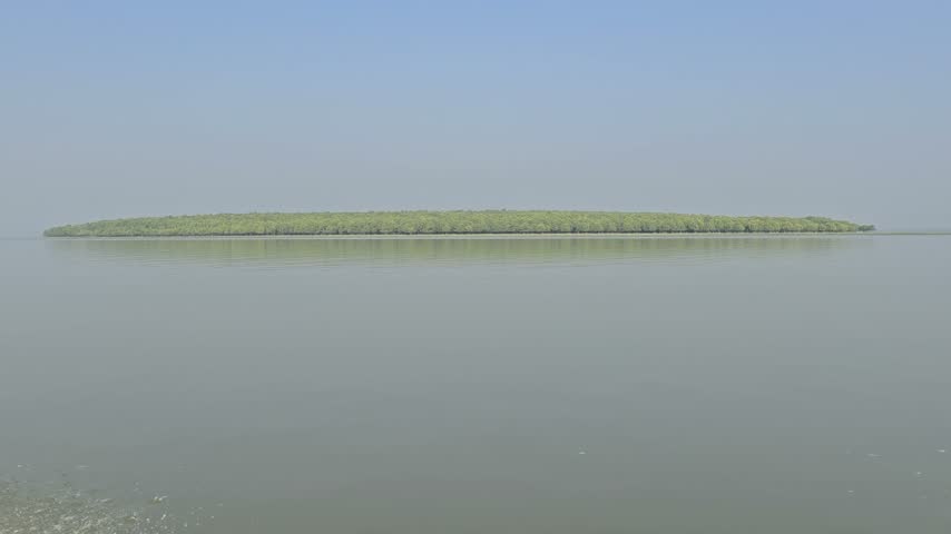 A wide panoramic view of a green island or landmass covered with trees situated in the middle of a calm sea or wide river under a clear blue sky." 