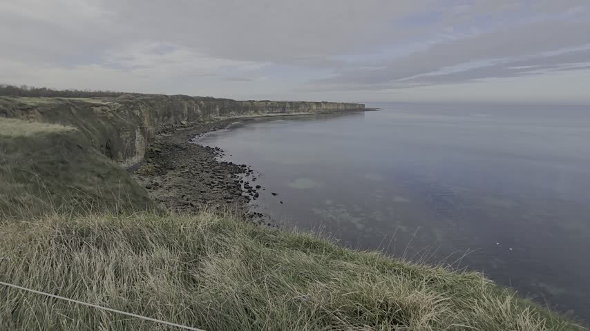 Shallow waves wash along a sandy shoreline beneath a layered, weathered coastal cliff.