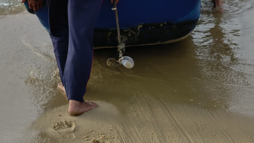 Barefoot local fisherman walking by a traditional wooden fishing boat with a propeller on a sandy beach in Central Vietnam