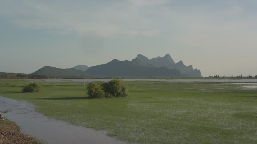 A tranquil view of a flooded grassy plain stretching toward a dramatic mountain range under a hazy sky during the late afternoon light