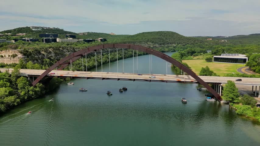 Wide high-angle aerial drone shot capturing the iconic weathered steel arch of the Percy V. Pennybacker Jr. Bridge (360 Bridge) crossing Lake Austin in Austin, Texas, with recreational pontoon boats cruising on the river below and the lush green Texas Hill Country cliffs in the background.