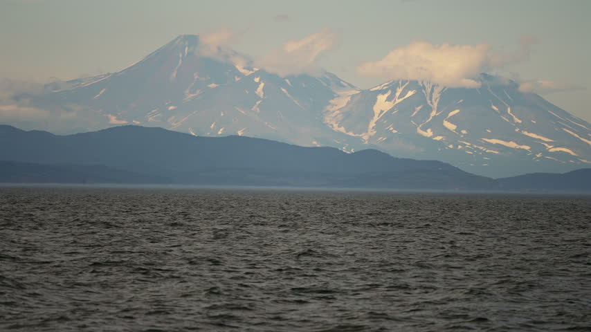 Slow motion wide view of humpback whale breaching in Pacific Ocean in front of two snow-capped volcanoes, Megaptera novaeangliae off coast of Kamchatka, Russian Far East