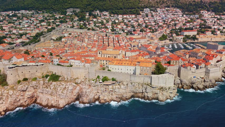 Dubrovnik Croatia fills aerial view with terracotta rooftops, historic stone walls and deep blue Adriatic water. Coastal old town rests beneath green hills as waves break along rocky shore.