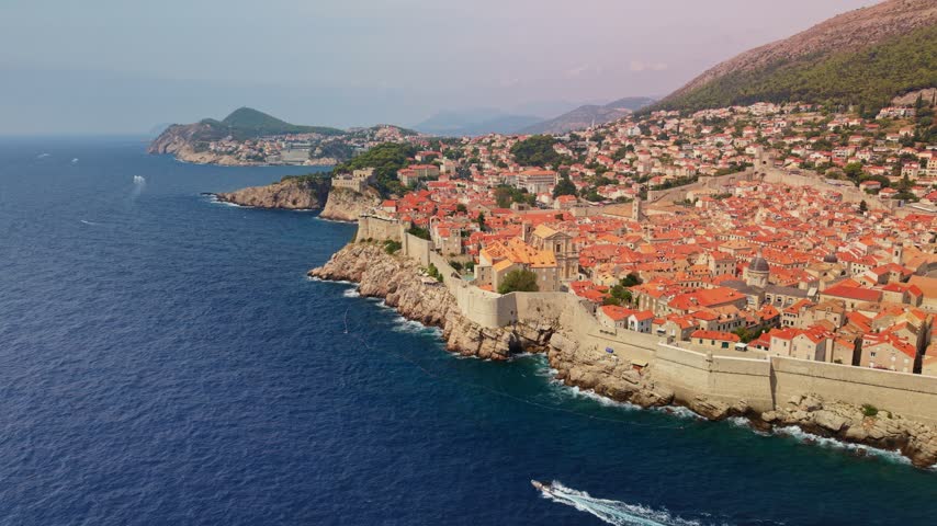 Dubrovnik Croatia stretches along coast, with pale stone fortifications and red tiled roofs overlooking Adriatic Sea. Offshore boat leaves long wake as homes climb distant hillside.