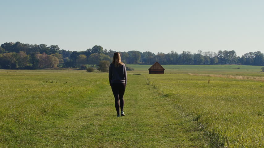 Woman walking through a scenic green meadow towards a traditional historic wooden hay barn in Trebon, Czech Republic, slow motion