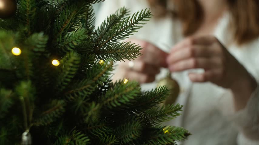 Woman decorating Christmas tree, hanging golden Christmas ornaments.