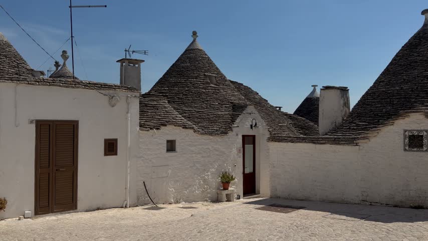 Slow forward view into a small sunlit square surrounded by whitewashed trulli in Alberobello, Puglia, Italy, with three different roof finials, an arched glass door and a tall white chimney