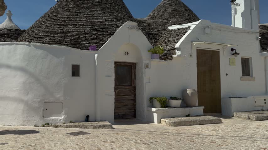 Whitewashed trullo facade with two weathered wooden doors and stone steps in Alberobello, Puglia, Italy, with conical roofs, a roof finial and potted plants under a blue sky