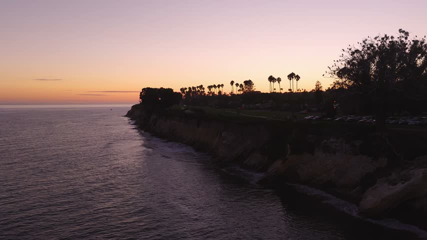 Aerial view gliding along the ocean bluffs of Santa Barbara, California during dusk, revealing silhouetted palm trees, seaside park paths, and rolling Pacific waves below.