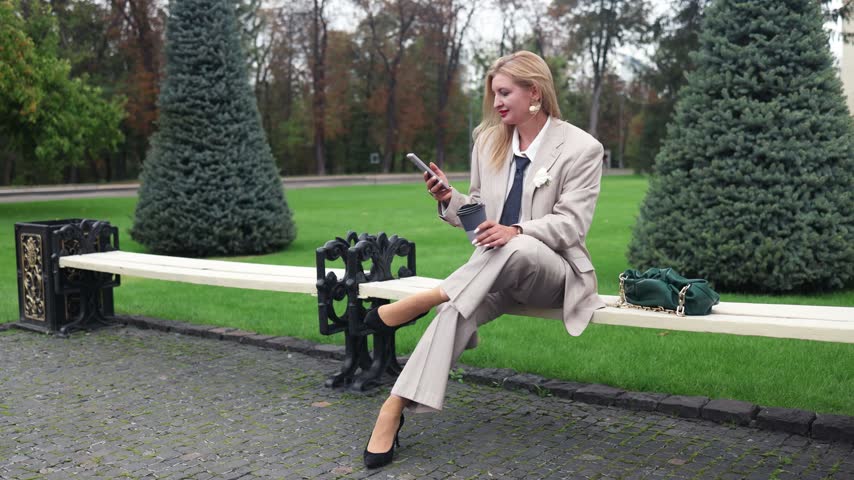 A stylish middle-aged blonde businesswoman with red lips wearing an elegant beige suit with a jacket, and high heels holding smartphone sits on a park bench during a coffee break.