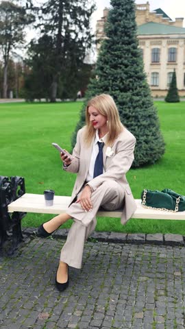 A stylish middle-aged blonde businesswoman with red lips wearing an elegant beige suit with a jacket, and high heels holding smartphone sits on a park bench during a coffee break.