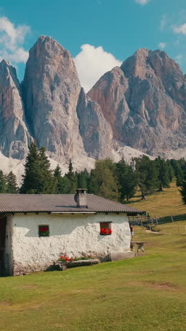 Alpine cabin under sharp Geisler mountain peaks in Dolomites. Traditional white hut stands on green grassy meadow near pine trees under blue sky with clouds in Italian alps in summer.