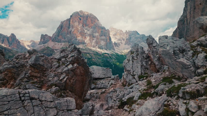 Camera moves forward between rocky cliffs toward Tofana peak in Italy. In motion reveals high stone mountain behind sharp grey boulders under cloudy sky in Dolomites region during summer.