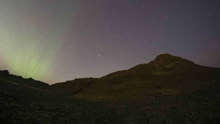 Serene starry night sky with glowing northern lights over dark rugged mountains and rocky terrain.   