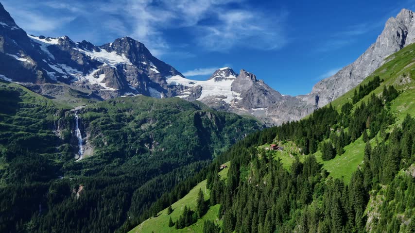 Drone view of a cascading waterfall, green alpine meadows, conifer forest and snow-covered mountain peaks under a blue sky.