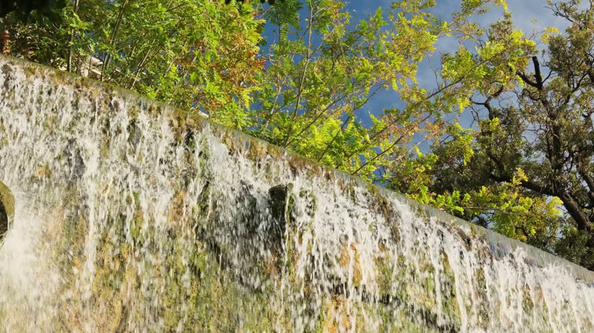 Low angle view of an artificial waterfall flowing down a stone wall, with lush green trees and a clear blue sky in the background, creating a serene and picturesque natural scene
