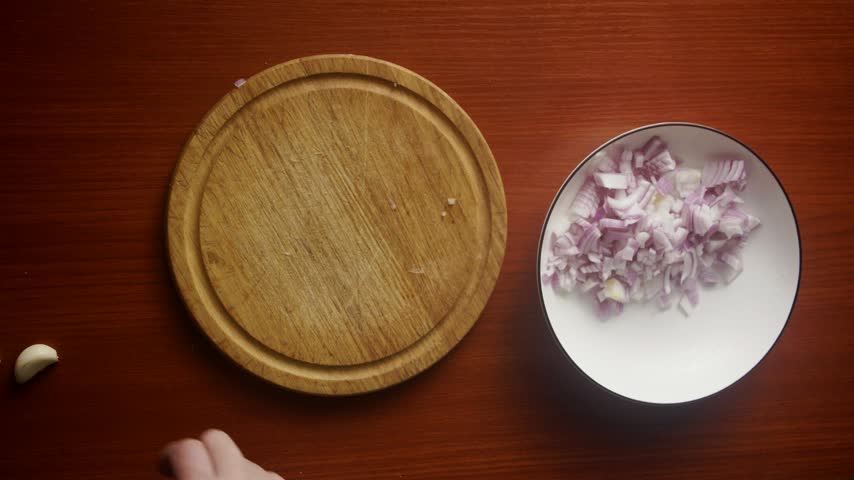 Horizontal Top-Down View of Hands Slicing Garlic Cloves next to Chopped Onions on Plate