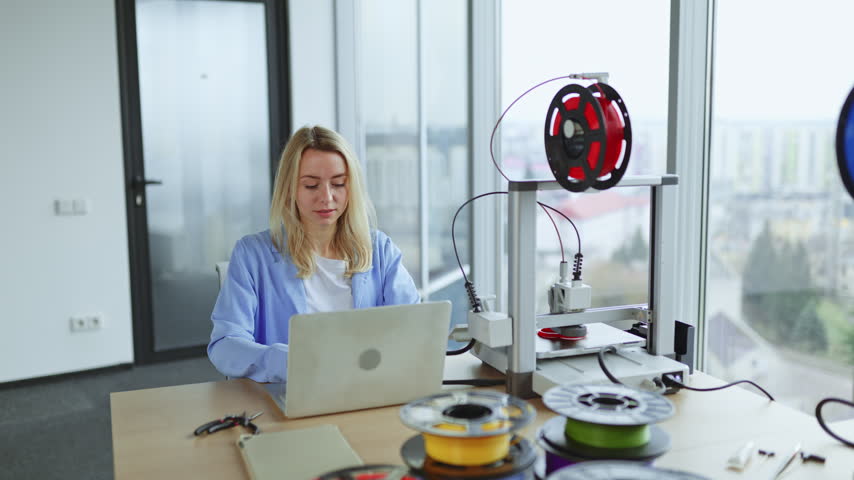 A jubilant woman celebrates while working with a 3D printer and laptop in a modern office setting with a city view