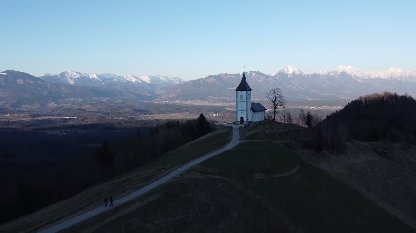 Aerial view of the iconic Jamnik church on a hilltop in Slovenia at dawn, with a winding path leading to its bell tower, silhouetted hikers, and the snow-capped Alps beyond.