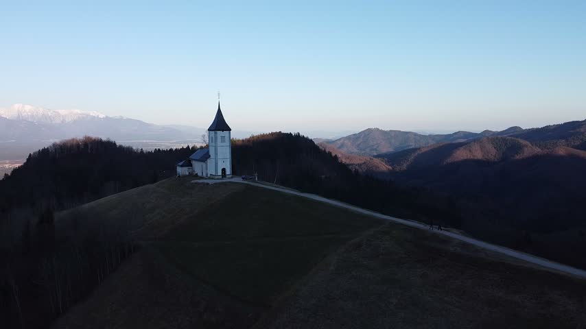 Aerial view of the iconic Jamnik church on a hilltop in Slovenia at dawn, with a winding path leading to its bell tower, silhouetted hikers, and the snow-capped Alps beyond.