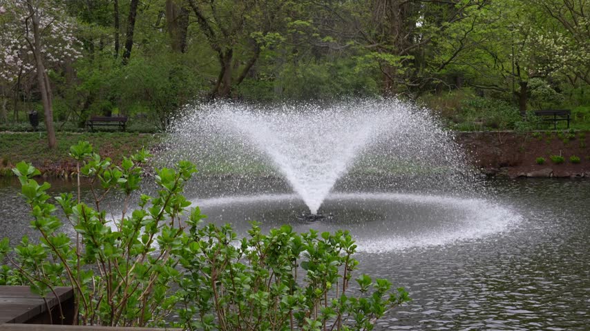Young boy in a blue jacket and striped hat walking beside a pond with a spraying fountain in a spring park. Concept of outdoor recreation, childhood exploration and a healthy lifestyle.