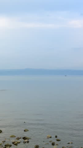 Peaceful evening view of a calm lake with distant mountains on the horizon and a small motorboat passing far away. A stone skips several times across the water surface, creating splashes and ripples in the tranquil lake scenery. Vertical nature video