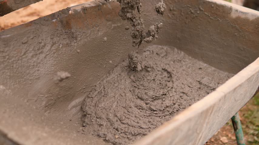 Wet gray concrete pours into metal wheelbarrow at outdoor construction worksite, building thick lumpy mound with rough damp texture under warm daylight in overhead view.