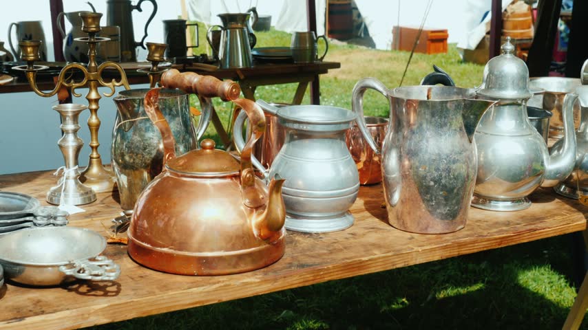 Close up view of antique style copper kettle pewter pitcher and silver candlestick arranged on rustic wooden table at historical marketplace outdoors under white canvas tents. Cinematic footage.