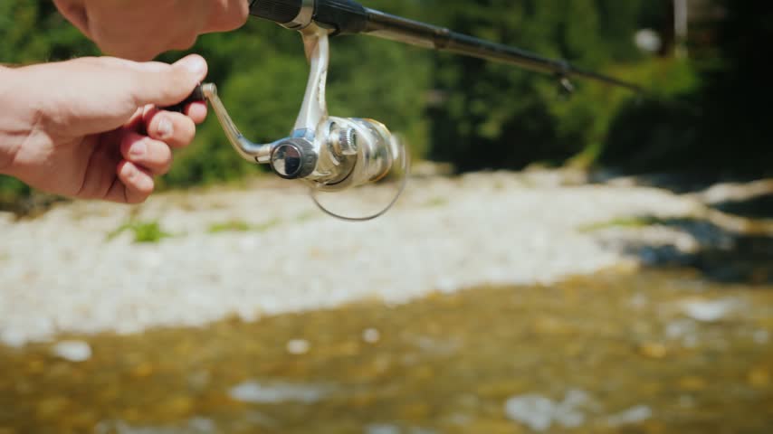 Close up shot showing hands winding fishing reel while spinning rod holds tension over shallow flowing rocky river water. Cinematic outdoor recreational activity footage.