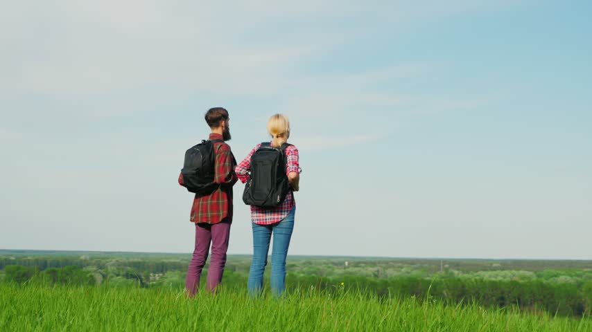 Rear view of young man and woman with backpacks standing on top of grassy hill looking at distant horizon, scenic rural landscape background during bright sunny summer day.