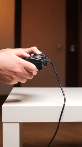 Vertical video hands gripping a black wired game controller on a white table, focused on casual indoor console gaming and leisure play