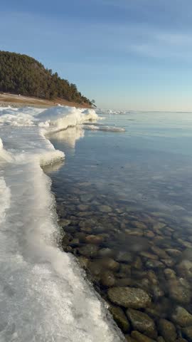 Vertical video of Baikal Lake on December morning. An ice-covered beach with calm blue water near village of Goloustnoye. Picturesque waterscape. Natural background. Travel and outdoor recreation