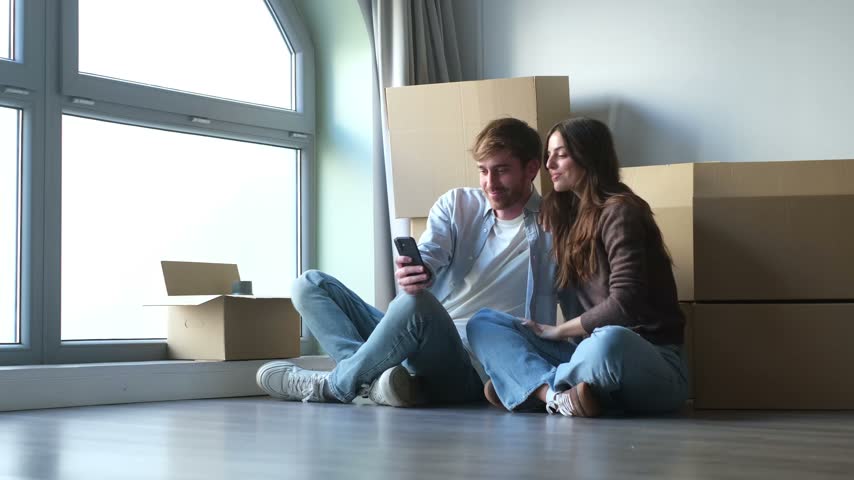 Couple sits on the floor in a new home, smiling and discussing content on a smartphone beside moving boxes. They share digital communication and online browsing while settling into their apartment.