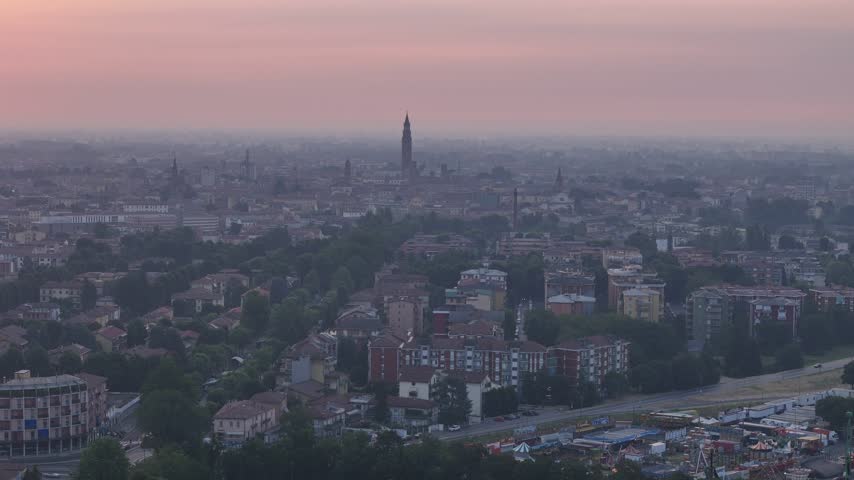 Cremona skyline rises around the Torrazzo bell tower in Lombardy, Italy, with dense urban districts, historic buildings and the Po Valley plain fading into morning haze, drone pullback.