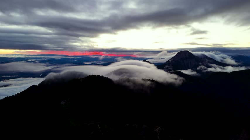 Vivid pink sunrise glow spanning wide horizon above vast sea of clouds blanketing valley, silhouetted Karawanks mountain peaks near Faaker See on Slovenia Austria border. Aerial drone wide shot